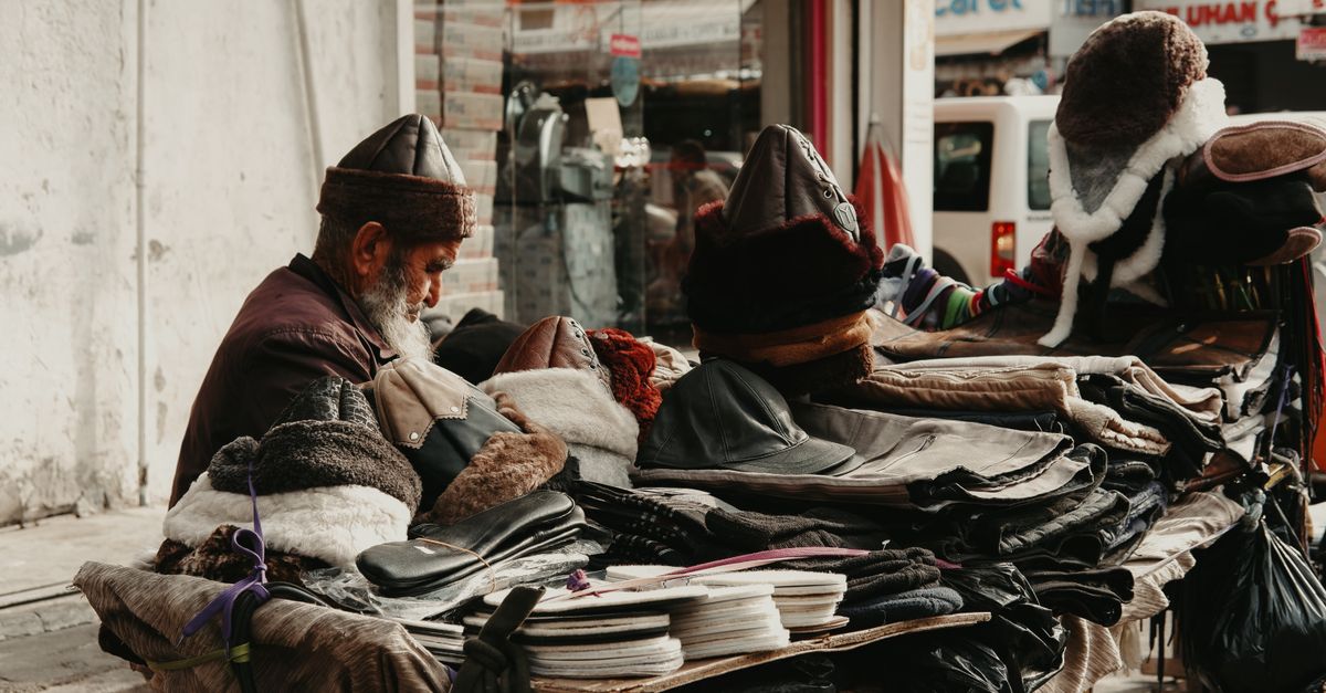 An elderly merchant selling handmade textiles and hats at an outdoor bazaar stall.